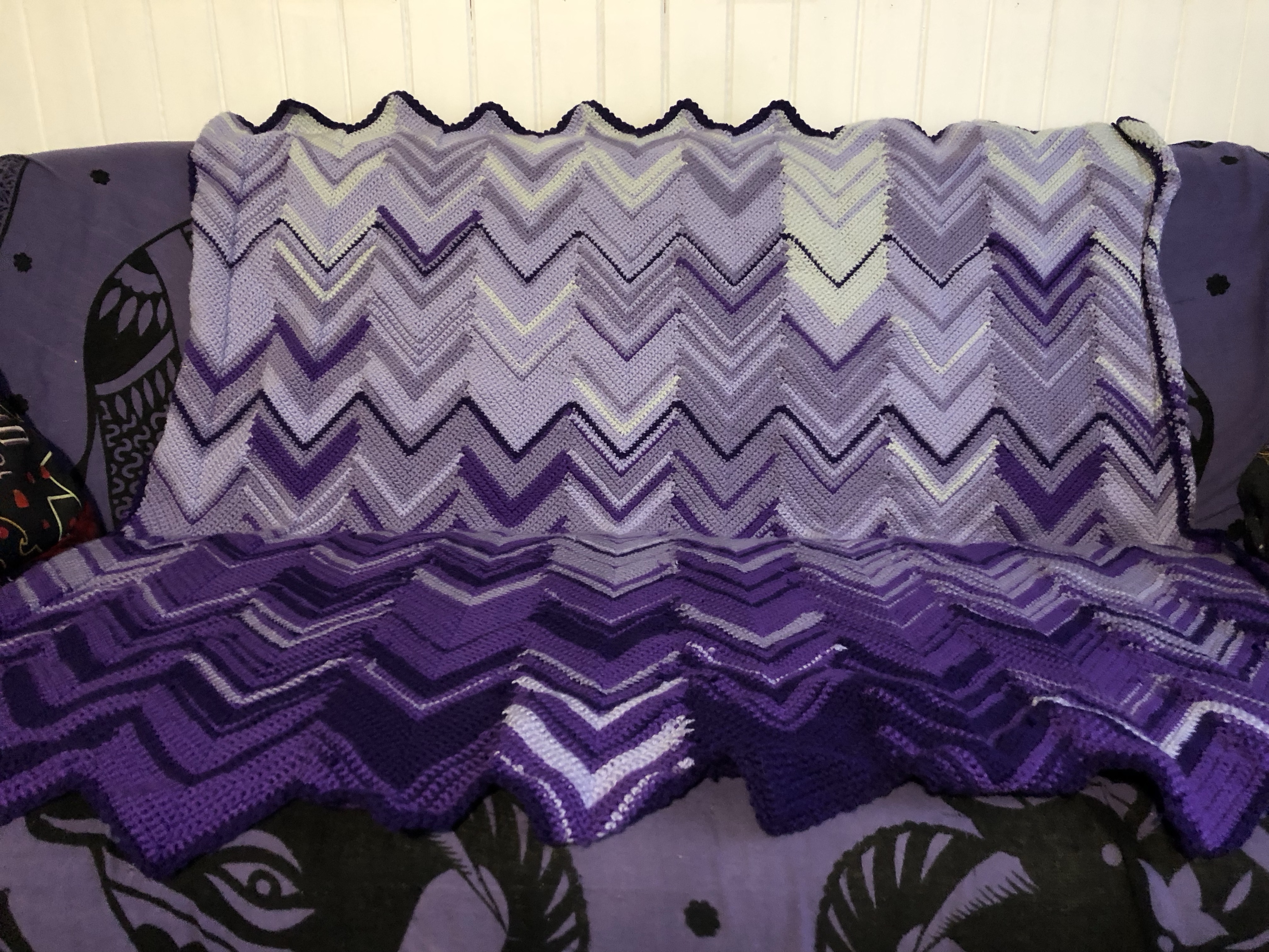 Blanket with a zigzag motif in different shades of purple, laid on a couch with a purple cloth cover
