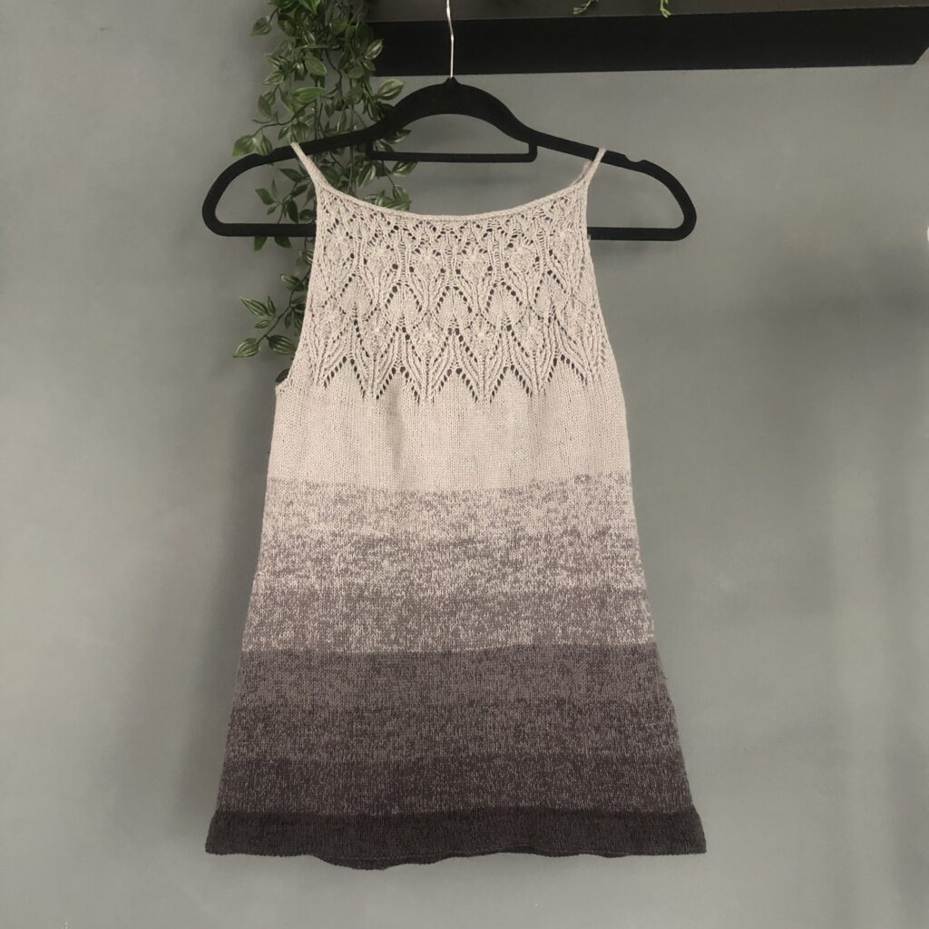 Tank top on a hanger, hanging from a black shelf, on a grey wall. Tank top is made with horizontal stripes going from a lighter grey on top to black on the bottom. 