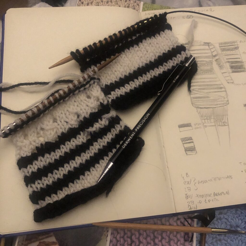 Two knitted rectangles with black and white stripes with different heights on needles on a notebook with a drawing of a striped minidress.