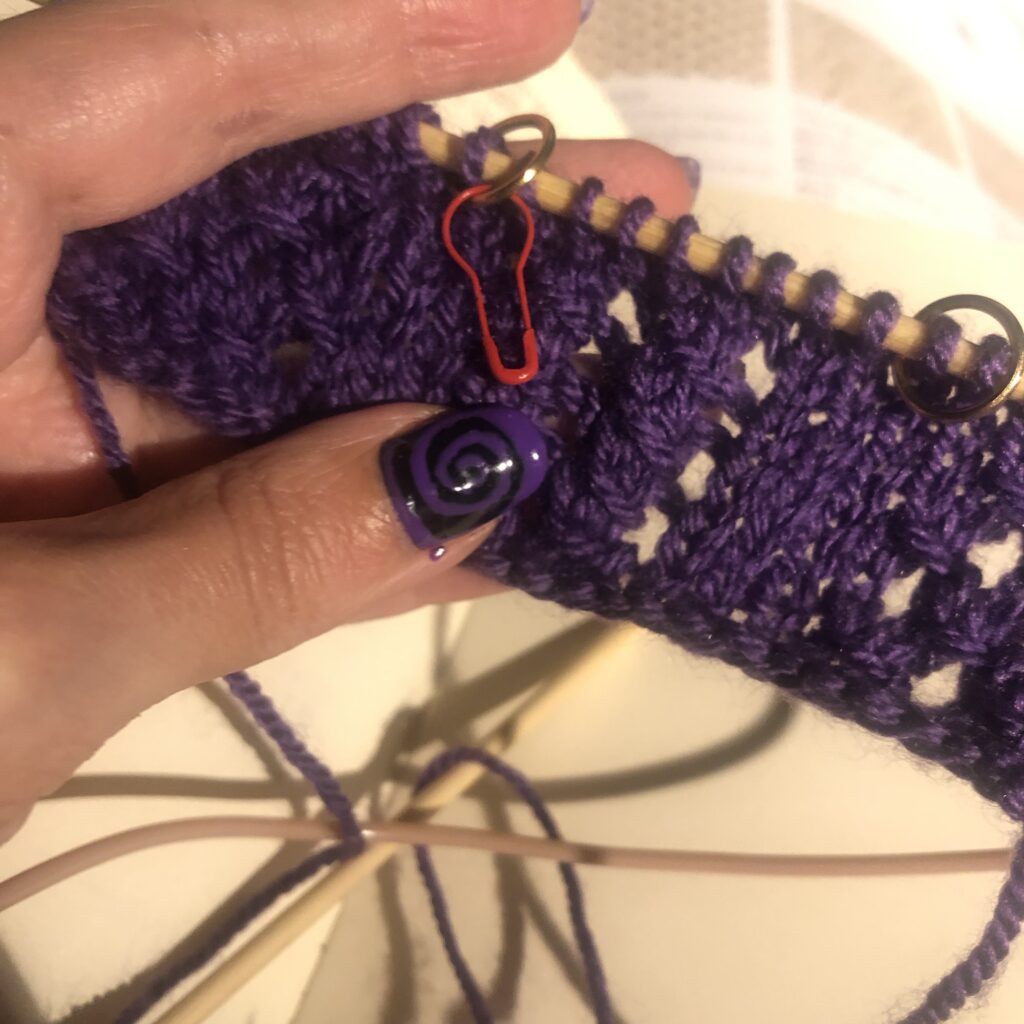 A purple knitted triangle on a needle held on a hand with a thumb on top with black nail and a purple spiral